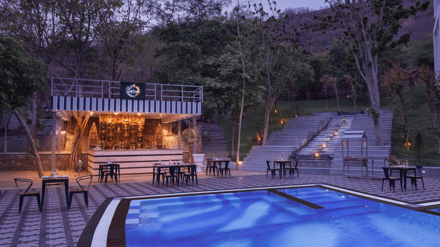 Outdoor poolside area at Essentia Luxury Resort & Spa, Udaipur, featuring a modern bar with seating, ambient lighting, surrounding trees, and steps leading up to a landscaped hill.