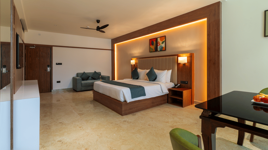 bedroom featuring a large bed with linens and a throw blanket, the room has light-colored marble flooring, wooden accents on the, a small round wooden table with two chairs, and armchair in the foreground at Essentia Resort & Spa, Sirsi 