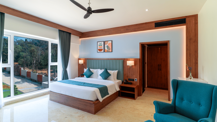 bedroom featuring a large bed with a dark wood headboard, bedside tables, a armchair in the foreground, a ceiling fan, and a large glass window offering a view of greenery outside at Essentia Resort & Spa, Sirsi 