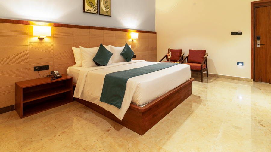 Bedroom featuring a large bed with linens and a bed runner, a dark wood bedside table with a lamp, and two chairs in the corner, the room has light-colored walls and tiled flooring at  Essentia Resort & Spa, Sirsi 