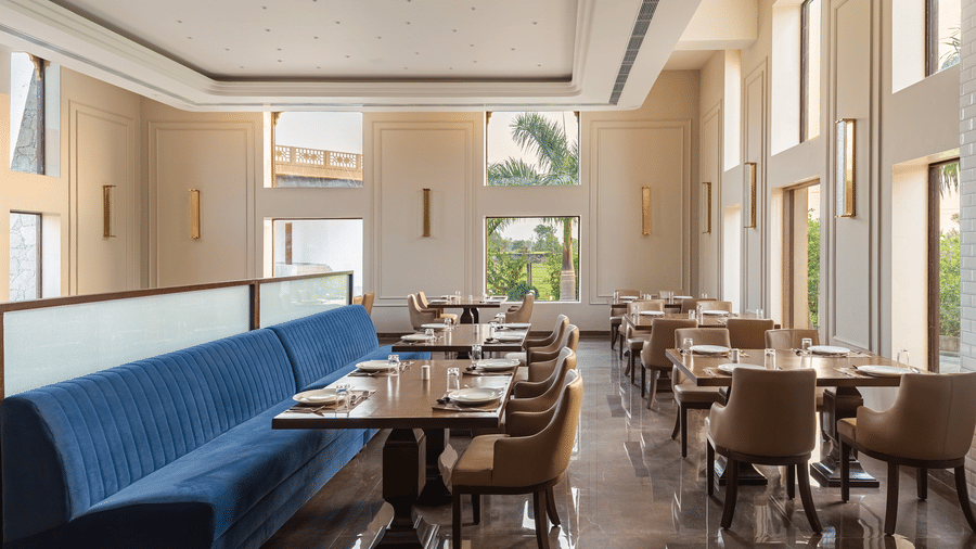 A view of the restaurant at EsthereaRaj Leela, Ranakpur, with long blue sofas, wooden tables, and neutral-coloured wall.