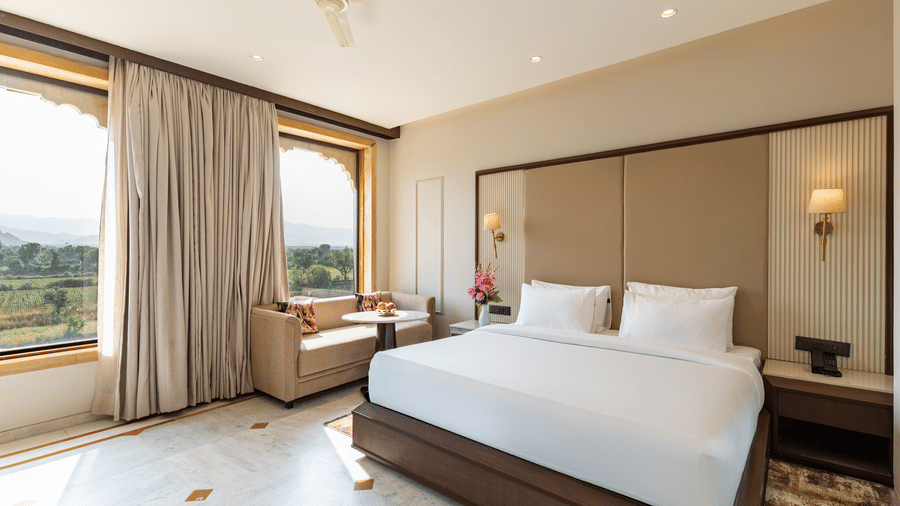 Bedroom at EsthereaRaj Leela, Ranakpur, featuring a king size bed, a ceiling fan, and a large window offering a scenic view of the green landscape.