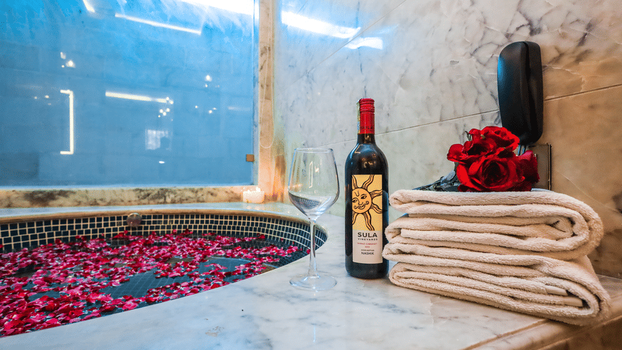 Detail of a bathtub at Estherea Resort & Spa, Jaipur with rose petals, a bottle of wine, and stacked white towels on a marble ledge.