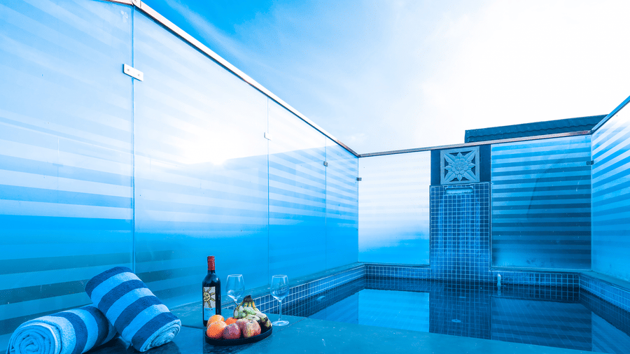 Rooftop pool area at Estherea Resort & Spa, Jaipur with frosted glass privacy panels, rolled blue towels, wine, and fruit by the water.