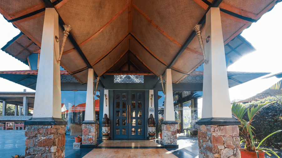 Entrance of dining at Estherea Resort & Spa, Jaipur featuring tall white pillars, stone bases, and a dark wooden door under a pitched roof.