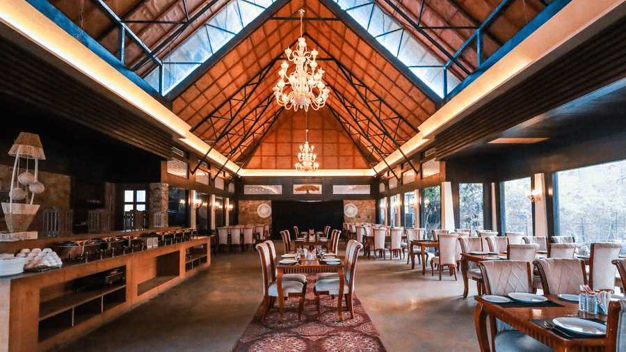 Wide view of a restaurant at Estherea Resort & Spa, a heritage resort near Jaipur showing rows of tables, a central aisle, and a high, patterned wooden ceiling.