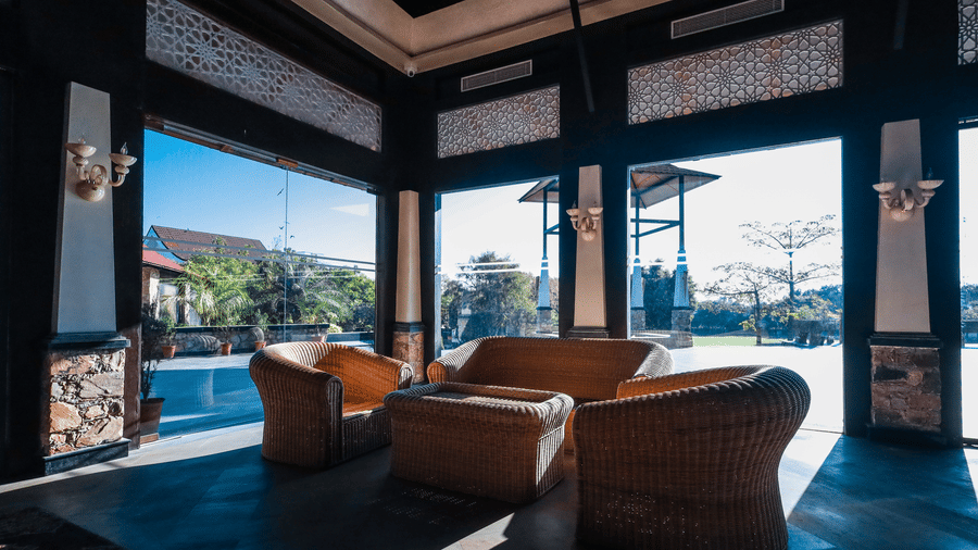 Semi-outdoor lounge at Estherea Resort & Spa, Jaipur with woven armchairs, a central table, and views of the garden through open arches.