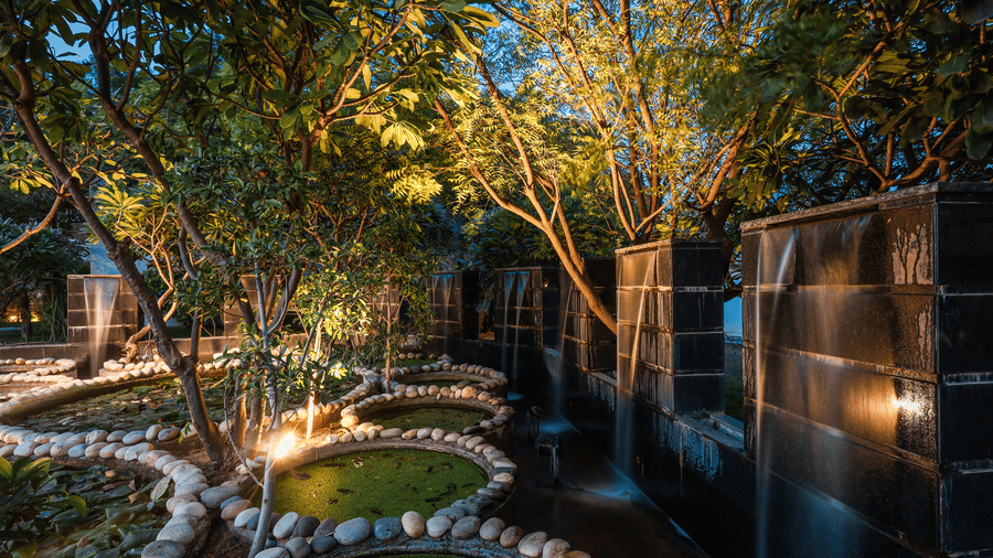 Evening view of the garden at Estherea Resort & Spa, Jaipur, with illuminated trees, stone ponds, and a modern water feature.