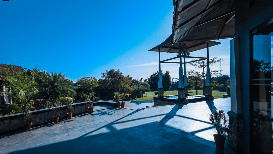An outdoor patio area at Estherea Resort & Spa, Jaipur with stone tiling, overlooking a green garden under a clear blue sky.