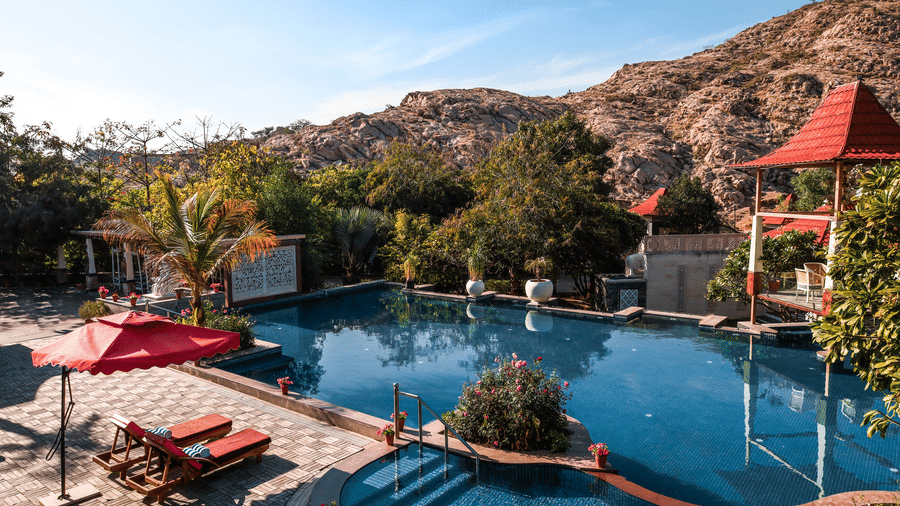 High-angle view of the outdoor pool at Estherea Resort & Spa, Jaipur, surrounded by red-roofed pavilions and mountainous landscape.