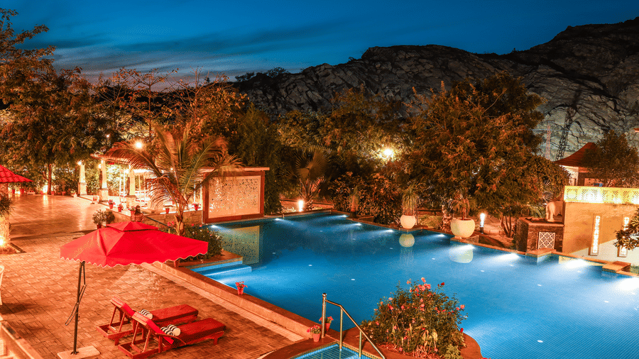 Evening view of the swimming pool at Estherea Resort & Spa, Jaipur, with red umbrellas and glowing underwater lights against a mountain backdrop.