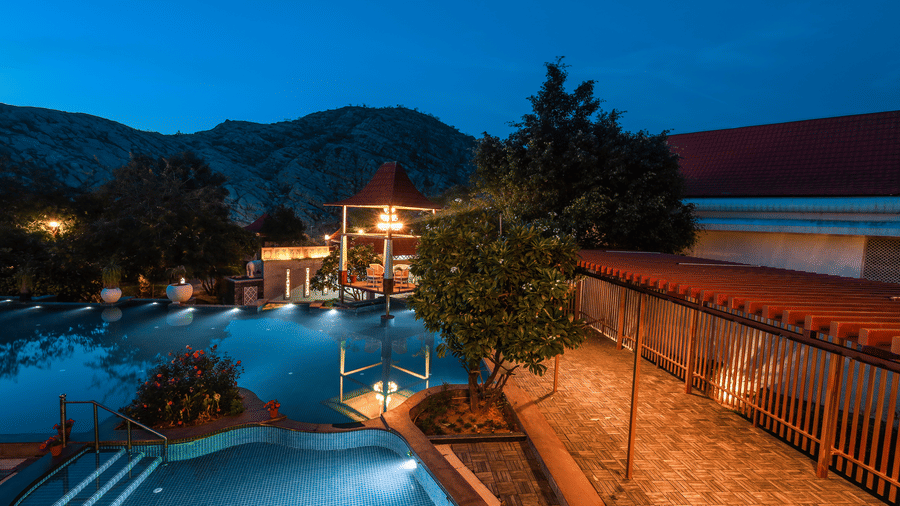 Nighttime view of the pool at Estherea Resort & Spa, Jaipur, featuring a lit fence and surrounding trees illuminated by soft garden lights.