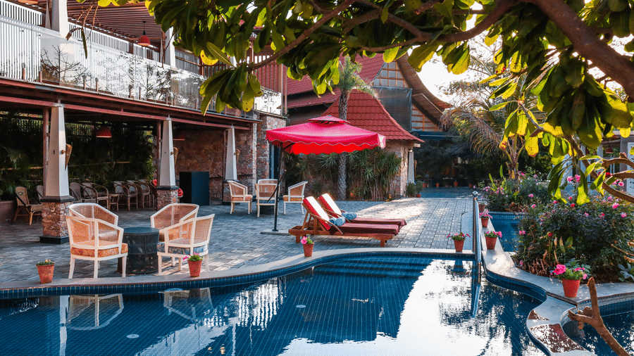 A curved outdoor swimming pool at one of the best luxury resorts near Jaipur, Estherea Resort & Spa, featuring wicker seating and red umbrellas on the surrounding stone deck.