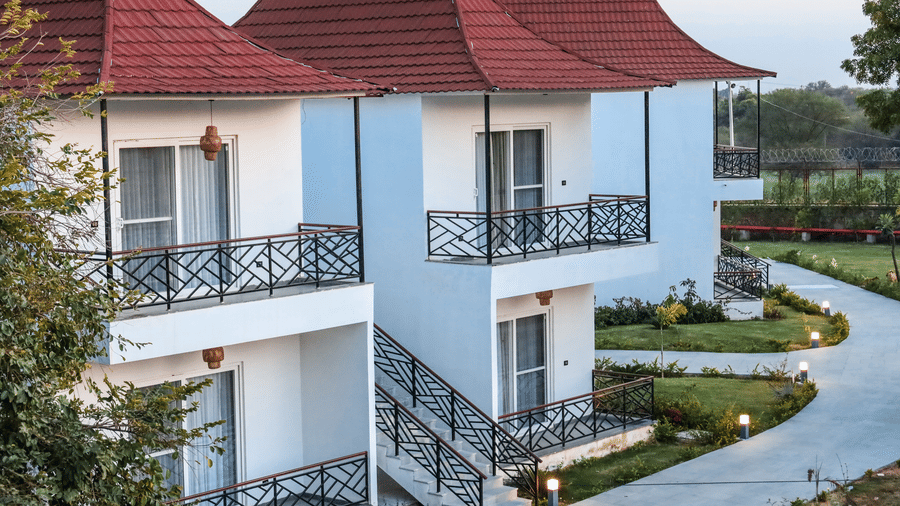 A row of white 2-storey villas at Estherea Resort & Spa, Jaipur with red pitched roofs, black metal balconies, and a paved walkway.