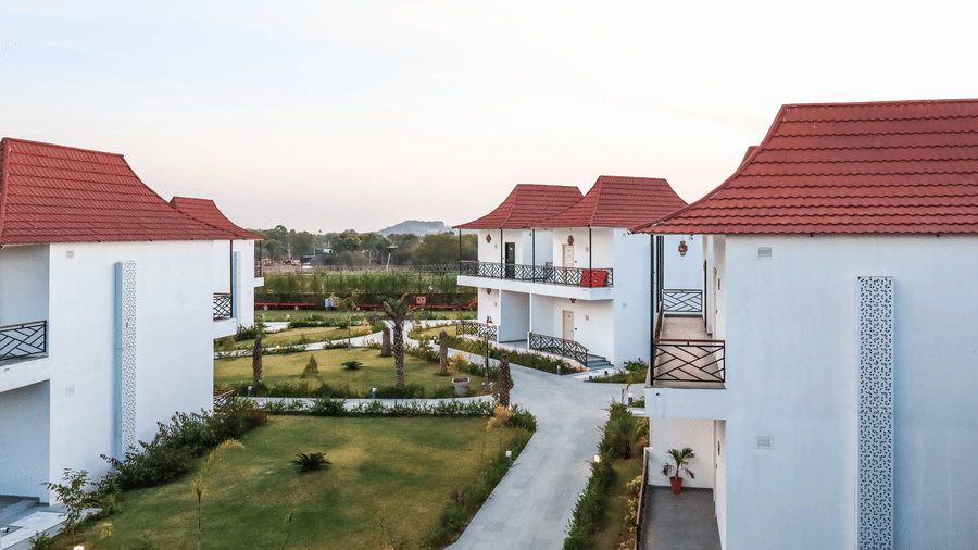 A row of white 2-storey villas at Estherea Resort & Spa, Jaipur with red pitched roofs, black metal balconies, and a paved walkway.