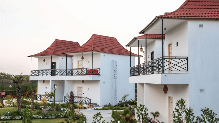 Multiple 2-storey white buildings at one of the best hotels with private plunge pools in room in Jaipur, featuring red peaked roofs, balconies with black railings, and a small garden path at Estherea Resort & Spa, Jaipur.