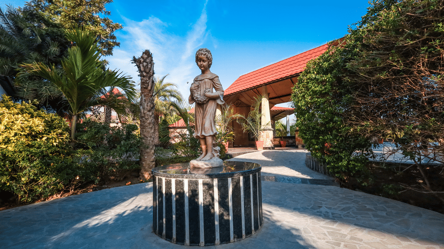 A bronze statue stands on a pedestal in a garden at Estherea Resort & Spa, Jaipur, with a sunlit path leading toward red-roofed structures.