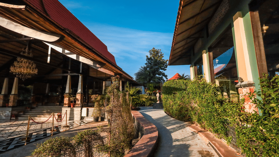Pathway lined with manicured hedges at Estherea Resort & Spa, Jaipur, leading past an open pavilion with dark wooden structural beams.