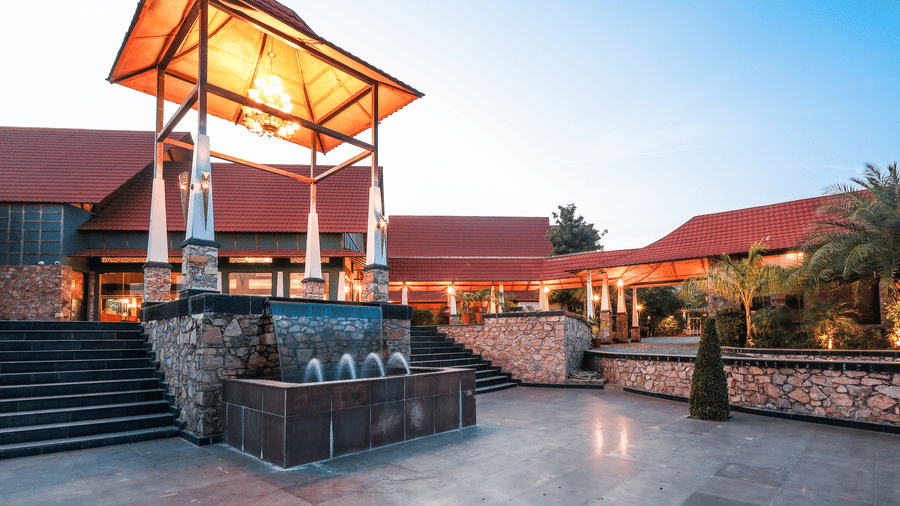 Stone staircase leads to a water feature and a lit tower at Estherea Resort & Spa, Jaipur, with red-roofed buildings in the background.