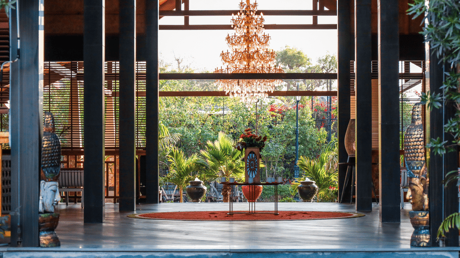 Open-air lobby at Estherea Resort & Spa, Jaipur, featuring dark wooden pillars, a central chandelier, and a view of the garden.