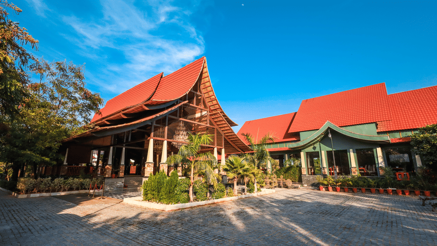 Red-tiled pavilion at Estherea Resort & Spa, one of the best resorts near Jaipur, featuring a steep roof, wooden pillars, and a stone-paved driveway under a clear blue sky.