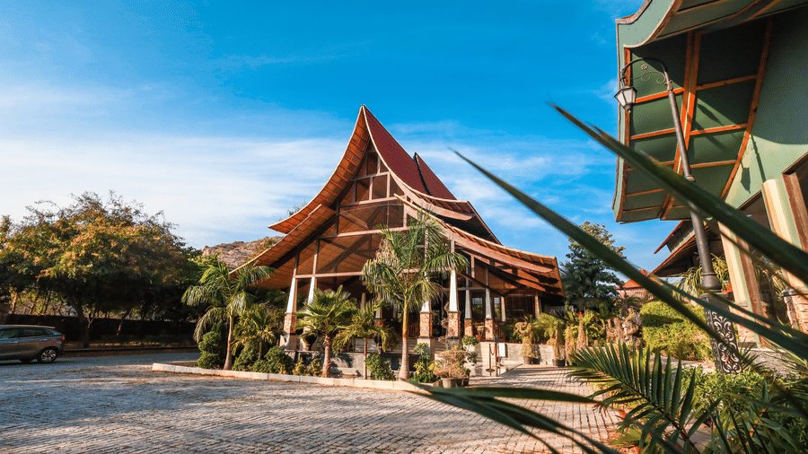 Perspective view of a traditional tiered roof building at Estherea Resort & Spa, Jaipur, surrounded by lush green trees and a paved road.