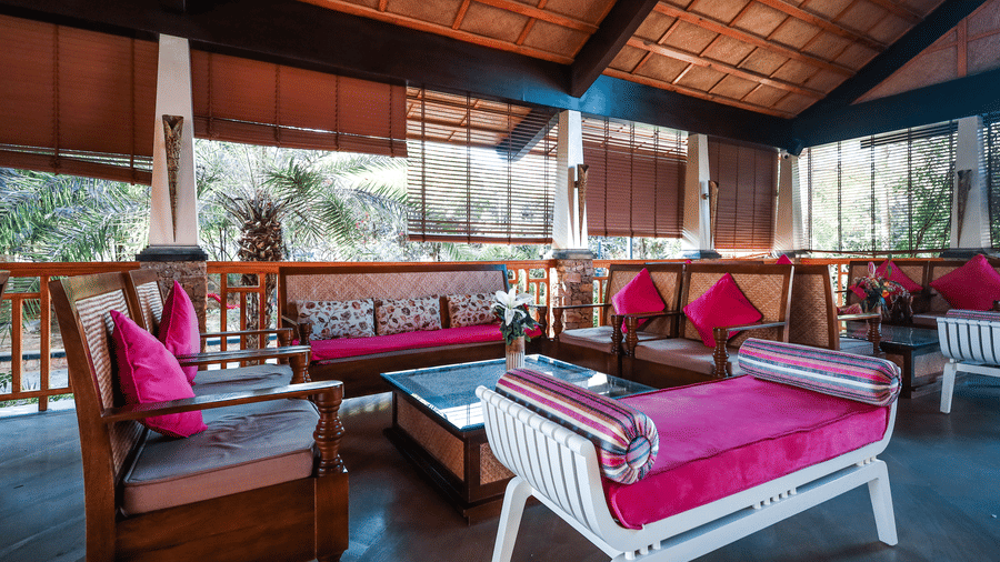 Indoor lounge at Estherea Resort & Spa, Jaipur, featuring wooden sofas with pink pillows and white benches under a high slanted ceiling.