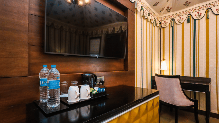Guest room interior at Estherea Resort & Spa, Jaipur, showing a wall-mounted TV, bottled water on a dark counter, and a desk with a chair.