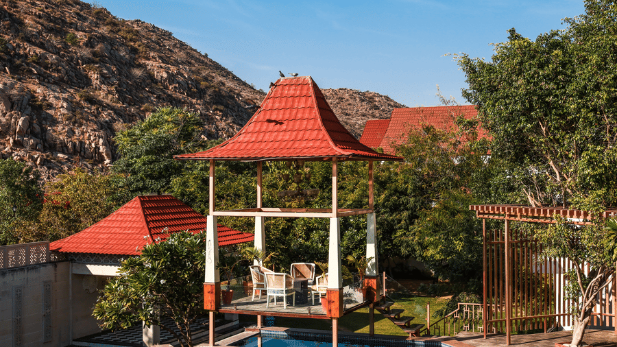 A red-roofed poolside pavilion at Estherea Resort & Spa, Jaipur, set against a backdrop of rocky hills and lush green trees.