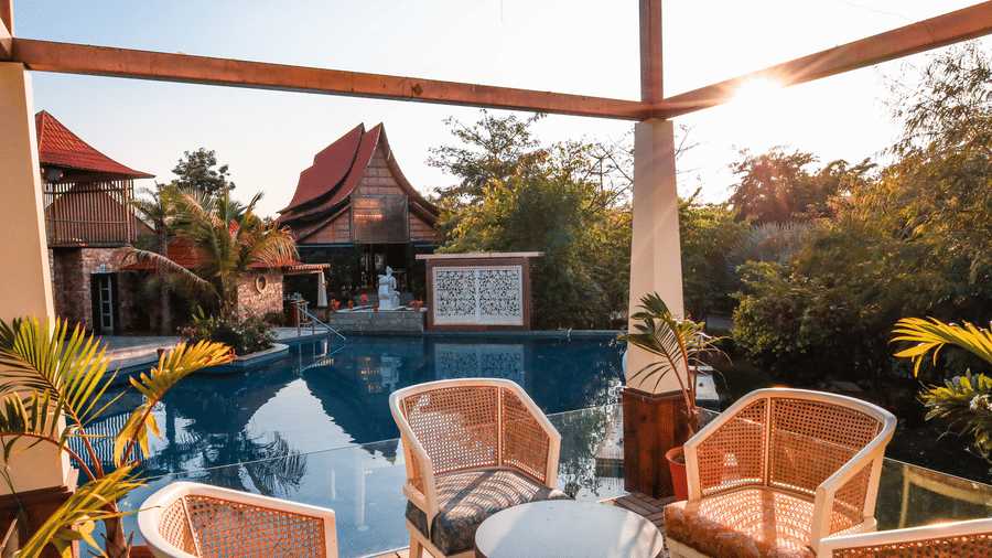 Wicker chairs and a table on a shaded terrace overlooking the swimming pool at Estherea Resort & Spa, Jaipur during a sunny afternoon.