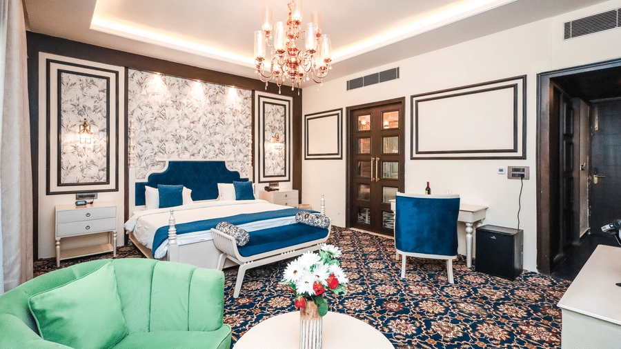 Bedroom at Estherea Resort & Spa, Jaipur featuring a blue upholstered headboard, patterned wallpaper, a chandelier, and a green sofa.