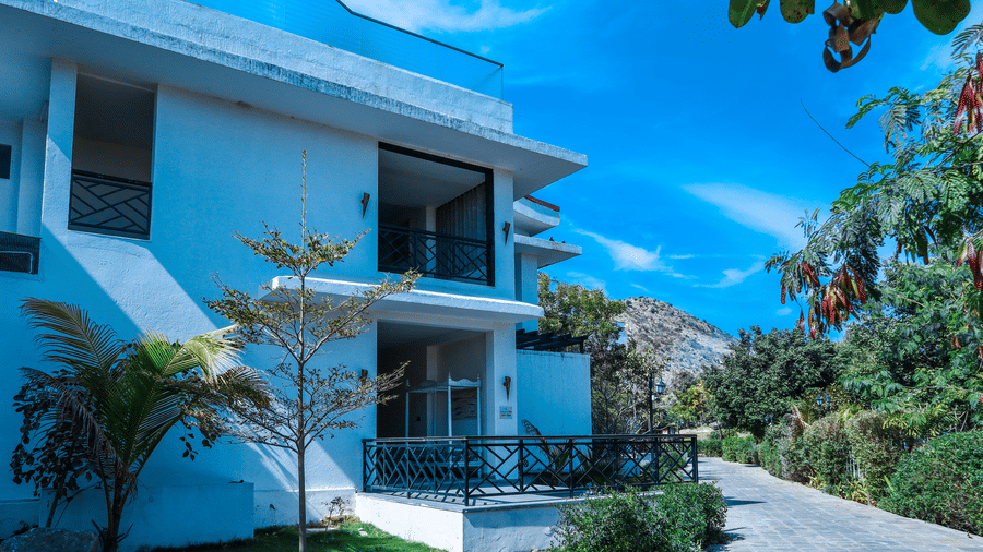 White multi-storey exterior building of Estherea Resort & Spa, Jaipur, showing balconies, green plants, and a paved walkway under a blue sky.