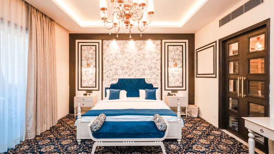 A suite with a blue velvet headboard, a chandelier, a patterned blue carpet, a white bench at the foot of the bed, and a large wooden door at Estherea Resorts & Spa, one of the best resorts near Jaipur with private pool.