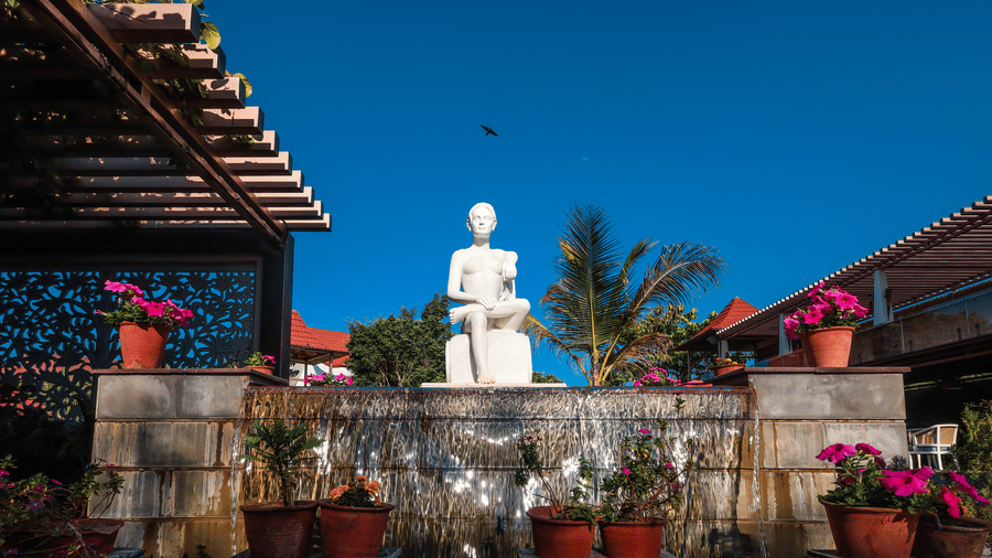 A white Buddha statue set against a stone water feature at the Estherea Resort & Spa, Jaipur pool area, decorated with pink flowers.