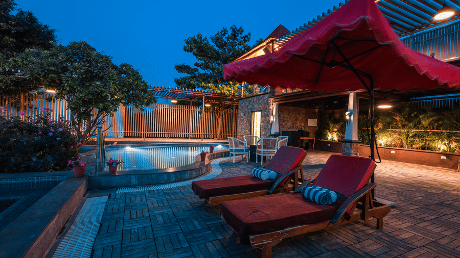 Poolside deck at Estherea Resort & Spa, Jaipur, featuring sun loungers and umbrellas next to the lit swimming pool.
