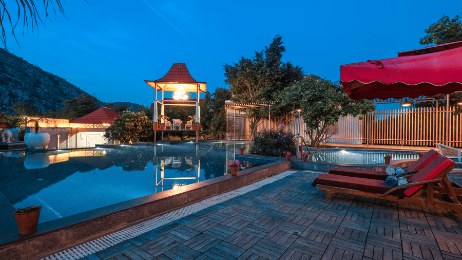 Night view of the swimming pool at Estherea Resort & Spa, Jaipur, with red lounge chairs on the deck and a lit pavilion in the background.