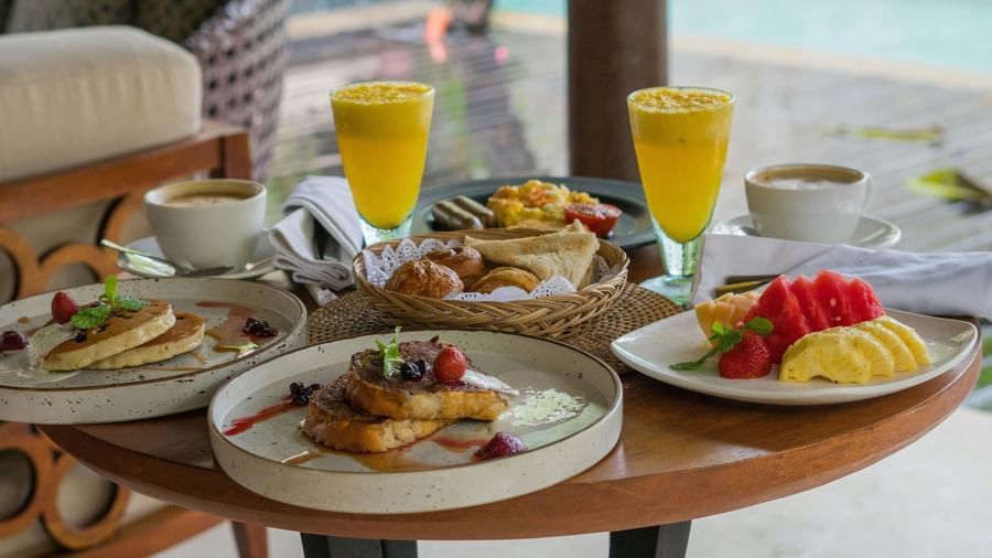 Round outdoor table with plates of pancakes, fruits, and glasses of fresh juice at Fireflies Resorts, Kabini.