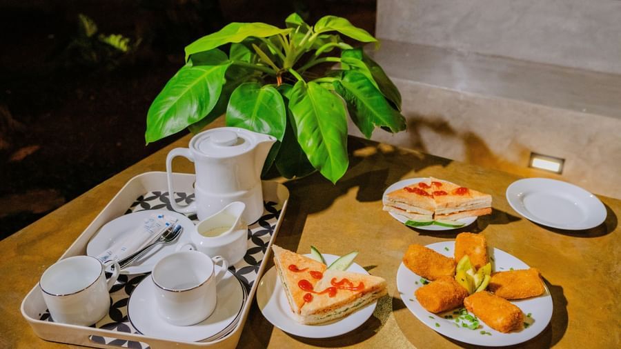 Tray with tea set and plates of sandwiches and fried snacks placed on an outdoor table near green foliage at Fireflies Resorts, Kabini.