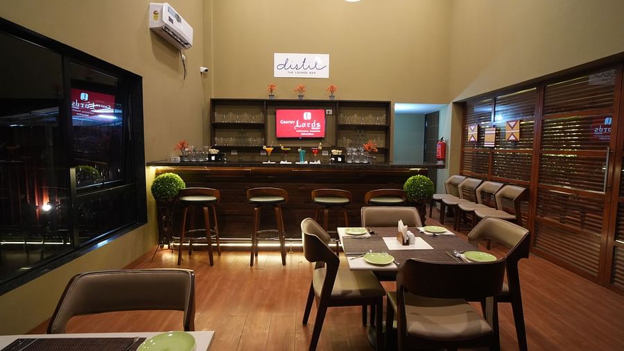 A cosy lounge bar with wooden flooring, high stools, cocktail displays, dining tables set with green plates, and large windows showing city lights at night - Gaurav Lords Resort Shrivardhan