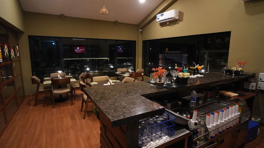 A cosy, modern restaurant bar with wooden flooring, neatly arranged dining tables, and a granite U-shaped counter stocked with glasses, syrups, and cocktail garnishes. Large windows offer a night view outside, and air conditioners are mounted on the walls 