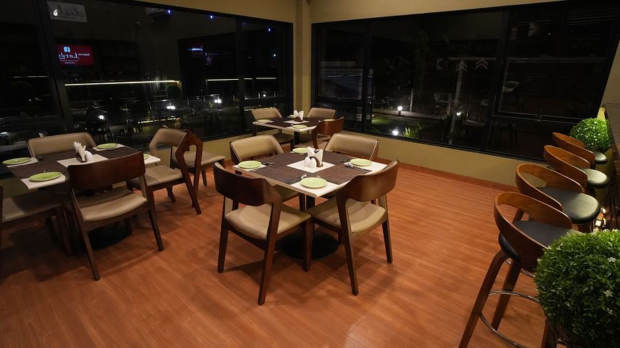 Indoor dining area with wooden flooring, cushioned chairs, neatly set tables, and floor-to-ceiling windows offering a night view, adjacent to a row of bar stools - Gaurav Lords Resort Shrivardhan