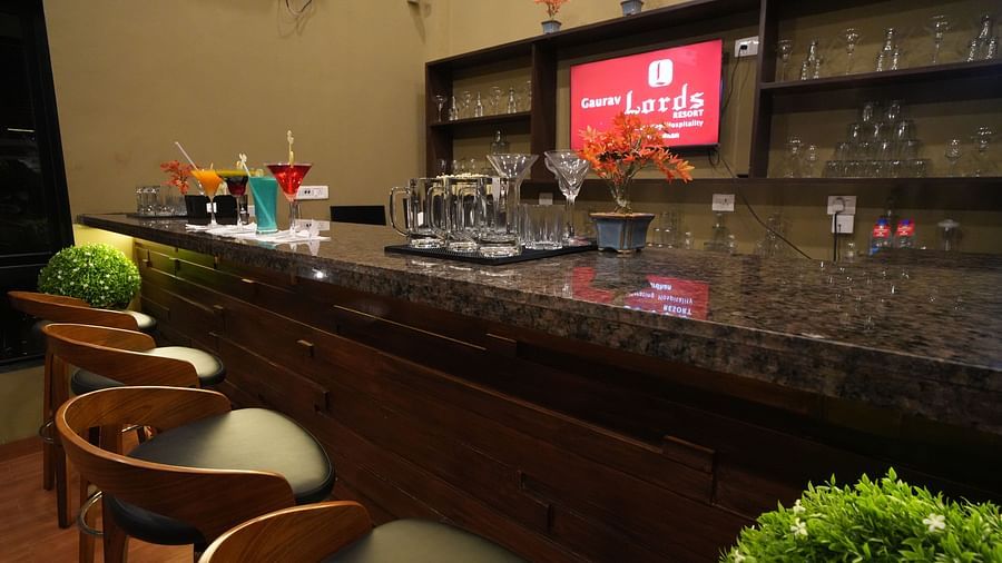 Close-up view of a modern bar setup with wooden high chairs, granite countertop, glassware, vibrant cocktails, decorative plants, and neatly arranged shelves in the background - Gaurav Lords Resort Shrivardhan