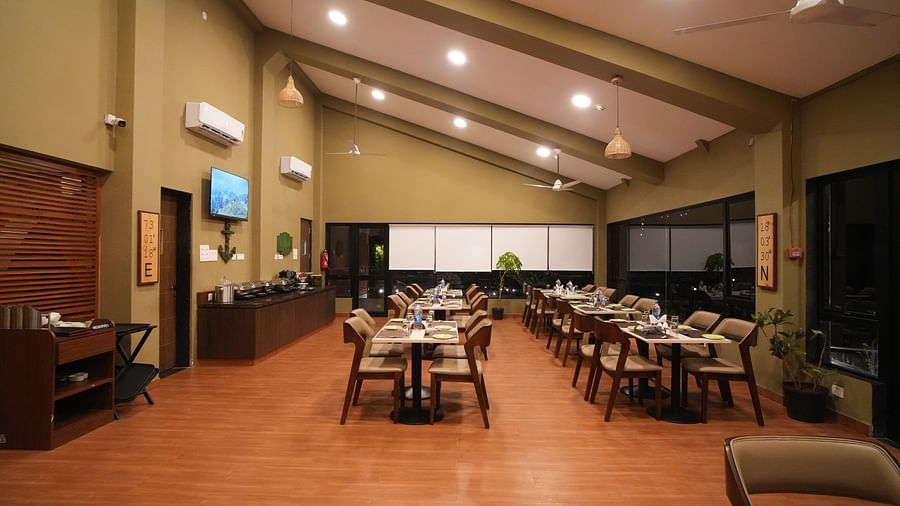 A restaurant with set tables and chairs, featuring a high, sloped ceiling and warm lighting. It includes a buffet area, air-conditioning units, and a wooden floor - Gaurav Lords Resort Shrivardhan.