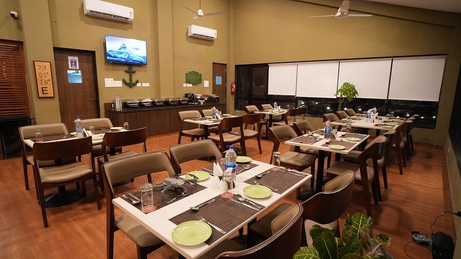 A restaurant with neatly arranged tables and chairs, offering a warm and inviting, dimly lit atmosphere. It features a television on the wall, air-conditioning units, and potted plants for added charm - Gaurav Lords Resort Shrivardhan.