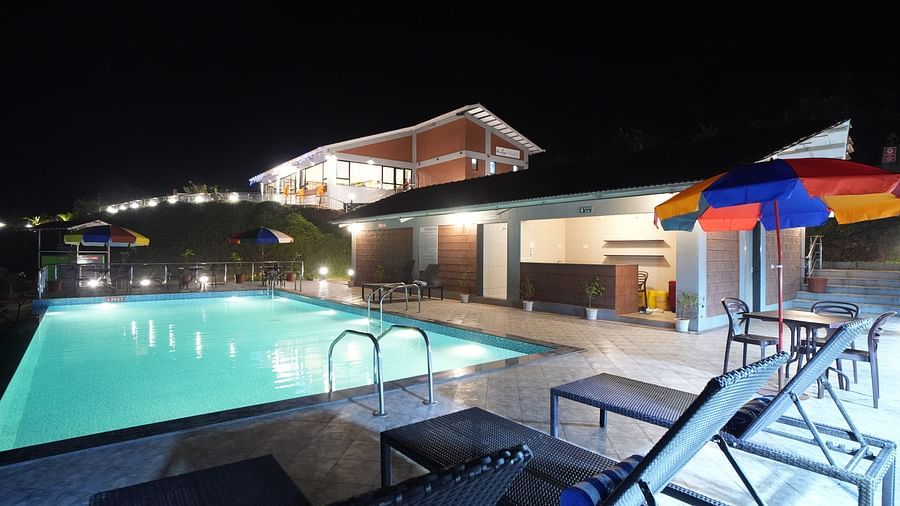 A nighttime view of a pool with lounge chairs in the foreground, illuminated by bright pool lighting, and buildings in the background. Colourful umbrellas are visible in the distance - Gaurav Lords Resort Shrivardhan.