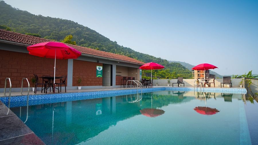 A swimming pool with red umbrellas and lounge chairs, set against a backdrop of mountains and a building - Gaurav Lords Resort Shrivardhan.