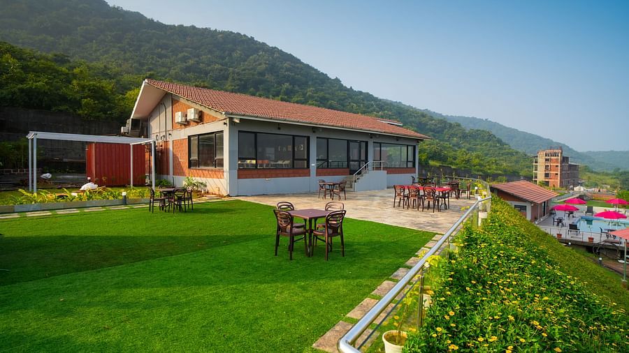 A building with a red roof and large windows, surrounded by a green lawn and a mountain backdrop, featuring outdoor tables and chairs and a metal railing in the foreground - Gaurav Lords Resort Shrivardhan.