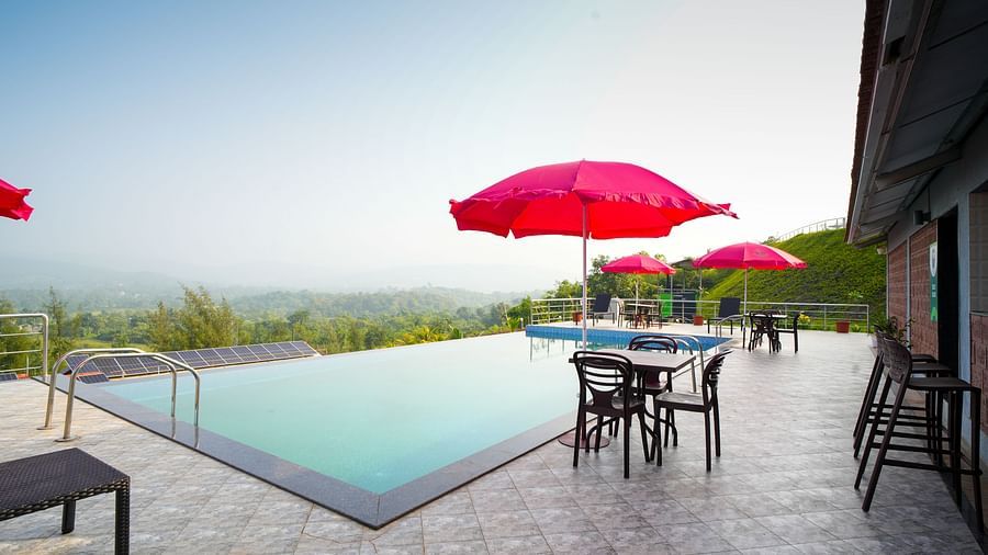 An infinity pool with red umbrellas and seating, offering views of a scenic landscape. It features a gray tiled patio and a building on the right side - Gaurav Lords Resort Shrivardhan.