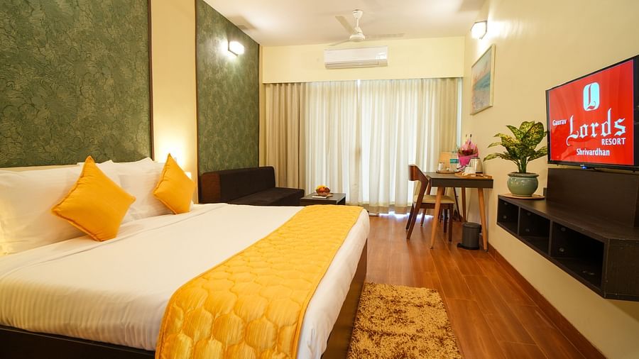 A hotel room with a bed, a television featuring the Lords logo, a dining table, a sofa, a rug, and a plant - Gaurav Lords Resort Shrivardhan.