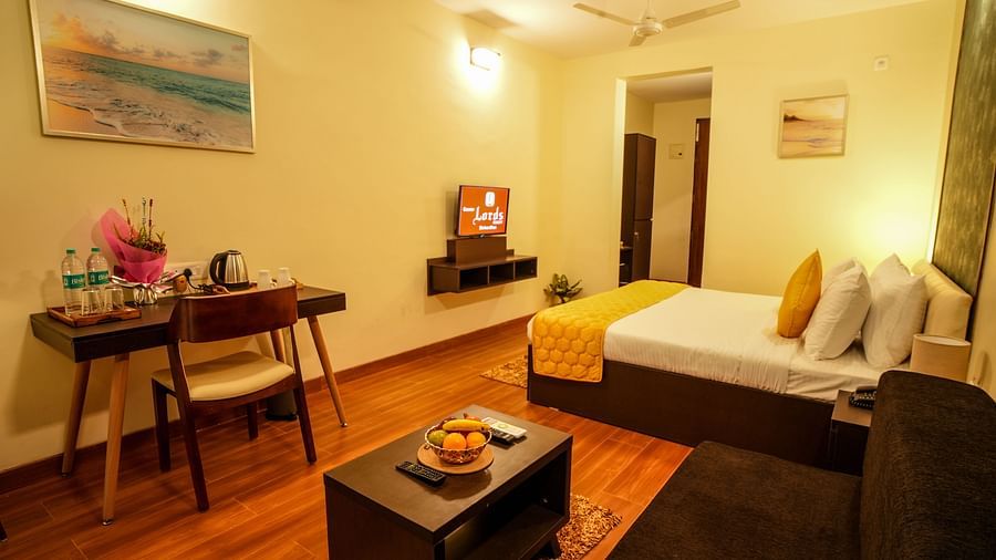 A hotel room with a king size bed, a work desk, and a small coffee table, featuring a flat-screen TV, a painting on the wall, and a doorway leading to another room - Gaurav Lords Resort Shrivardhan.
